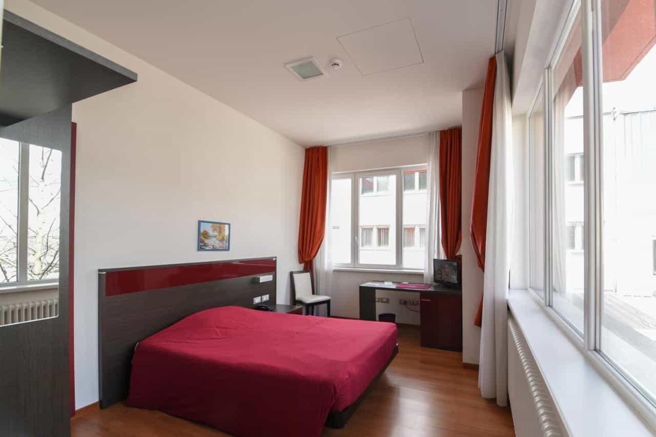 Hotel room with red bedding, wooden floor, and large windows with orange curtains.