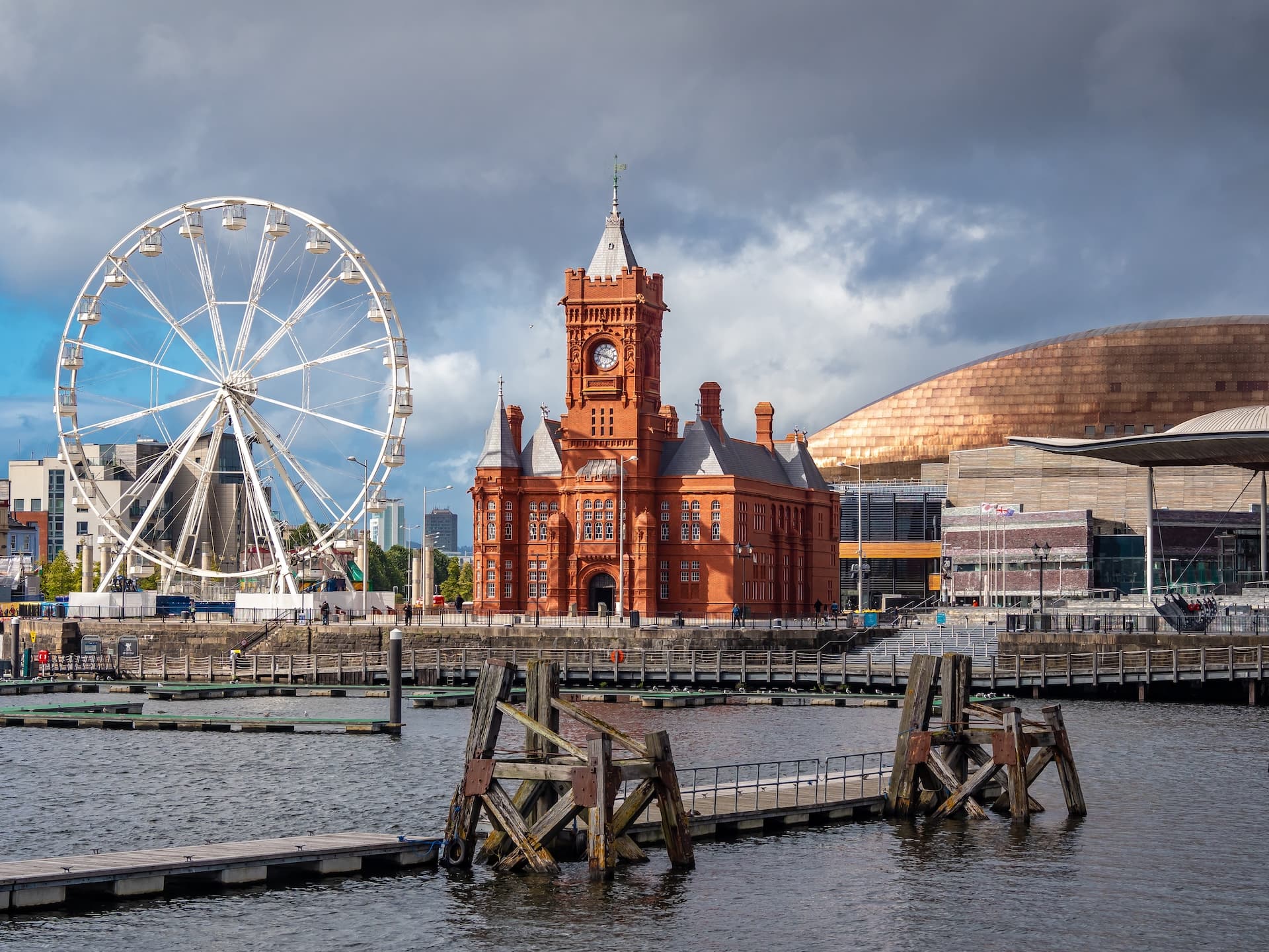 Ferris wheel and red brick clock tower by the water in Cardiff Bay under cloudy sky.