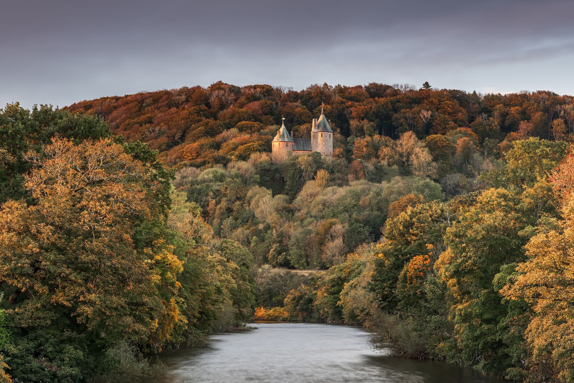 Castell Coch castle nestled in autumn foliage above a river valley under a gray sky.