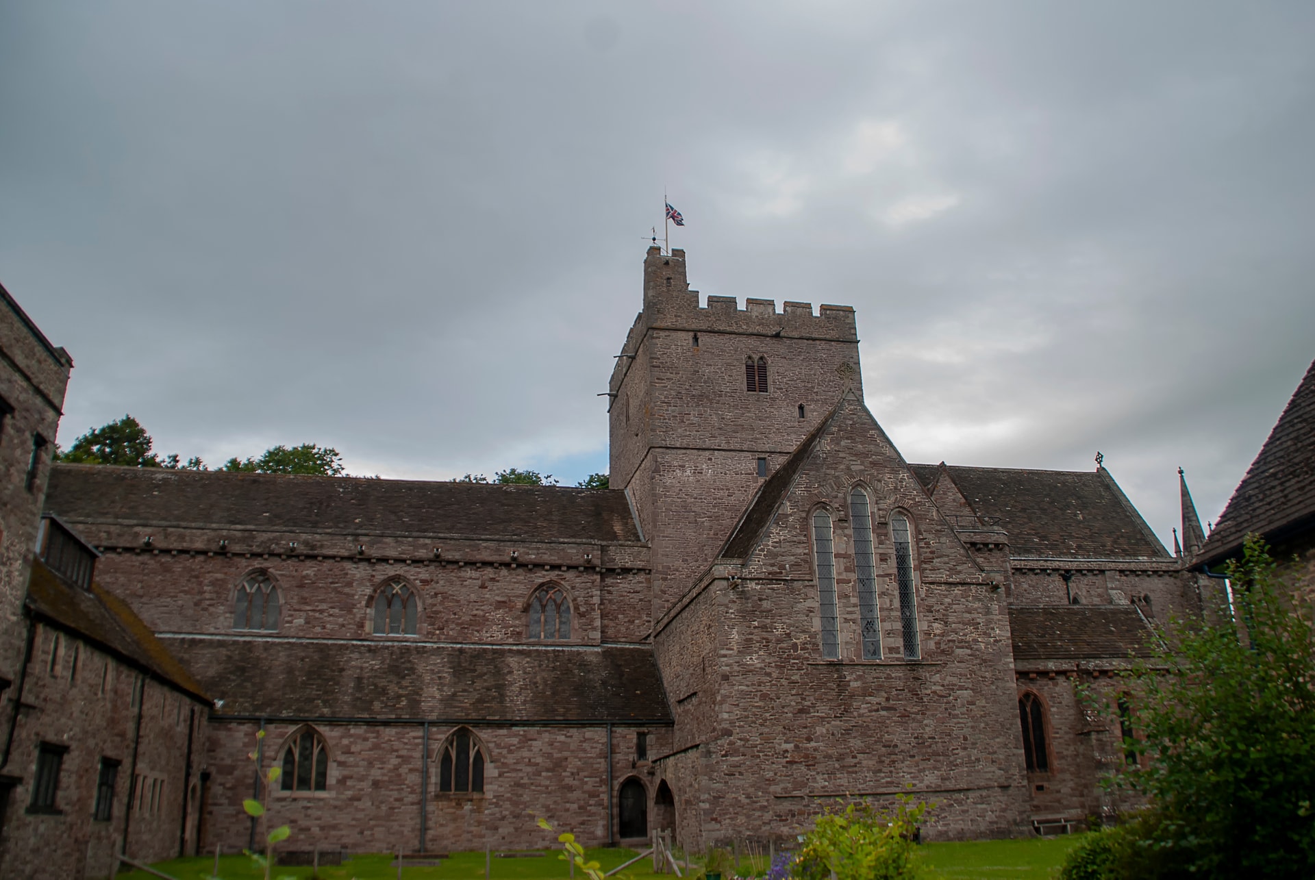 Brecon Cathedral exterior with stone walls and tower under a cloudy sky, Union Jack flying.
