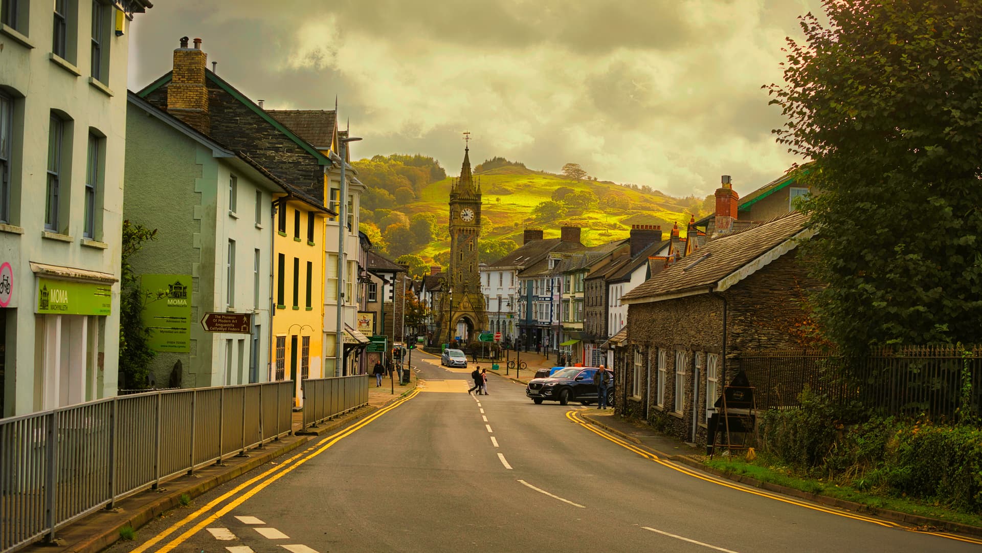 Street view in Machynlleth toward a clock tower with green hills under a dramatic sky.