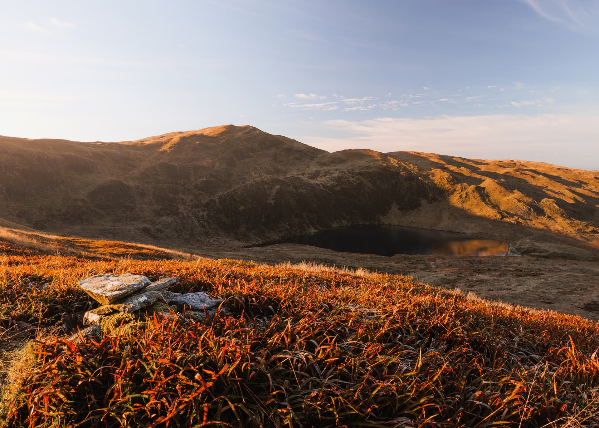 Mountain lake surrounded by brown hillsides at sunrise, with orange foreground grass and rocks.