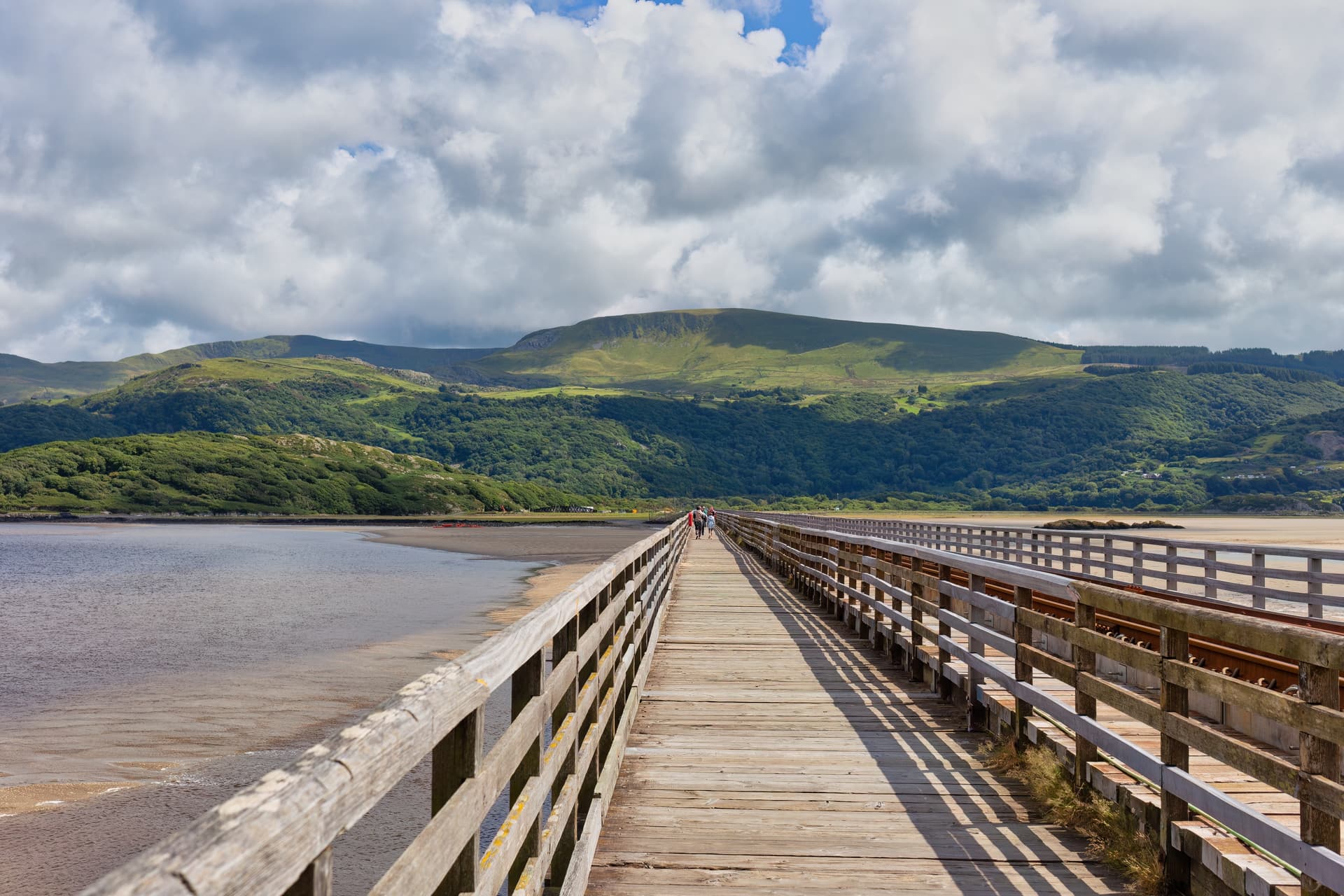 Wooden Barmouth Bridge walkway extending over water toward green hills under a cloudy sky.