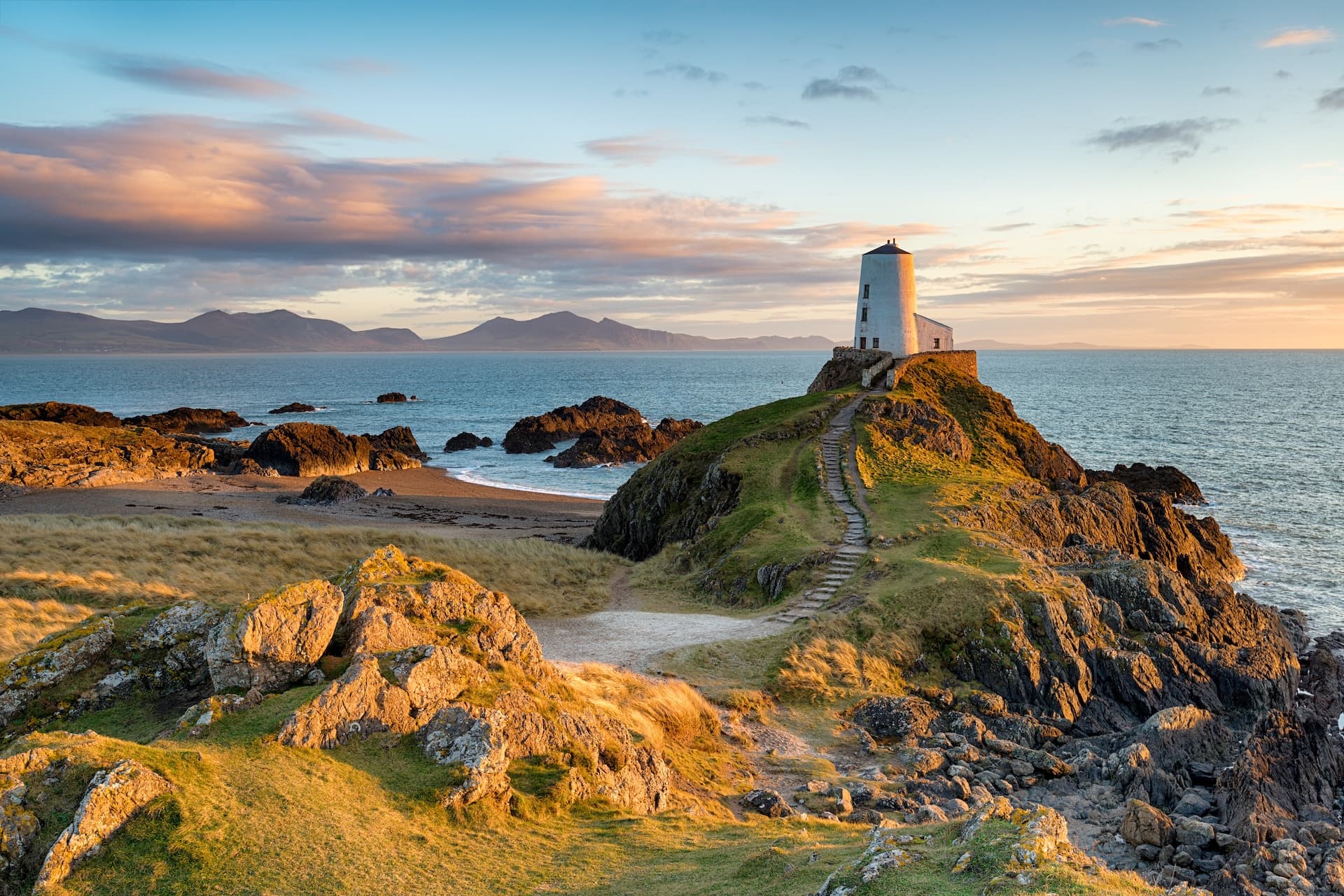 White lighthouse on rocky outcrop overlooking the sea with mountains in Anglesey at sunset