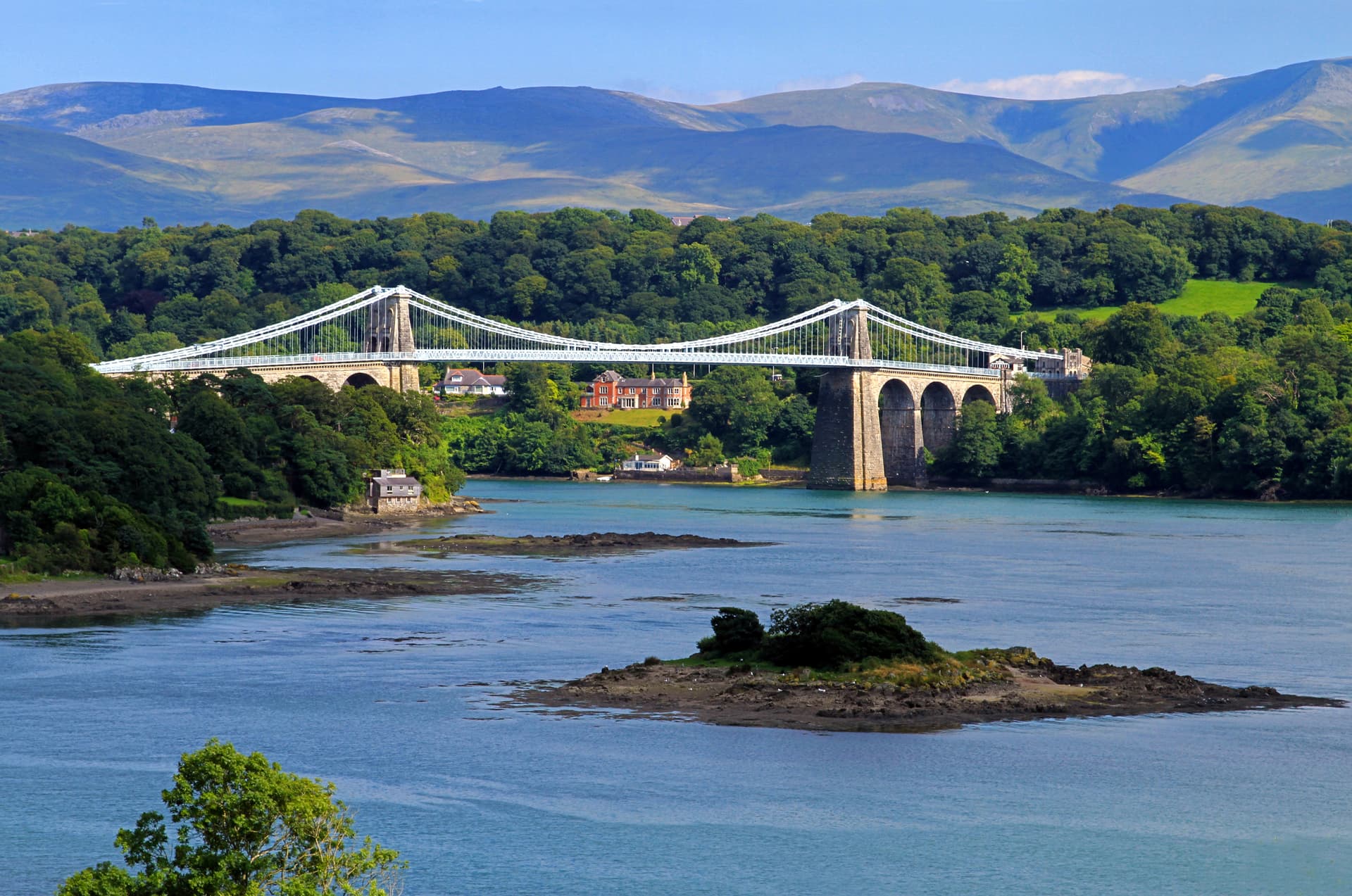 Suspension bridge over water with forested banks and mountains in the background, Menai Bridge.