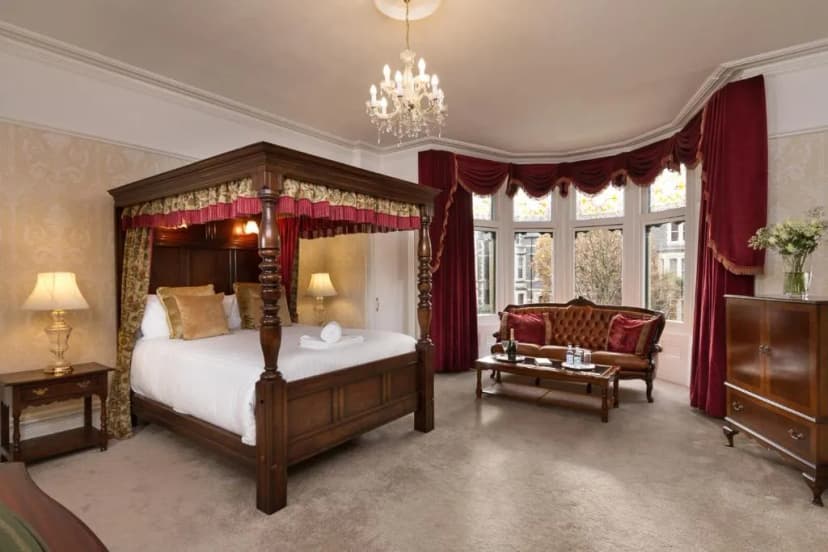 Four-poster bed and leather sofa in a hotel room with bay windows and red drapes