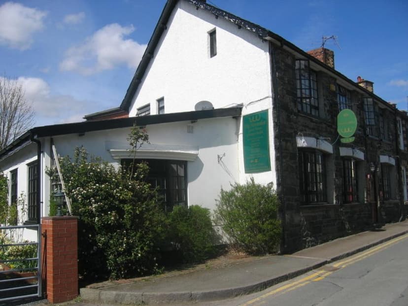 The Horseshoe Guesthouse with white and dark stone facade, green signs, and flowering bushes beside a street.