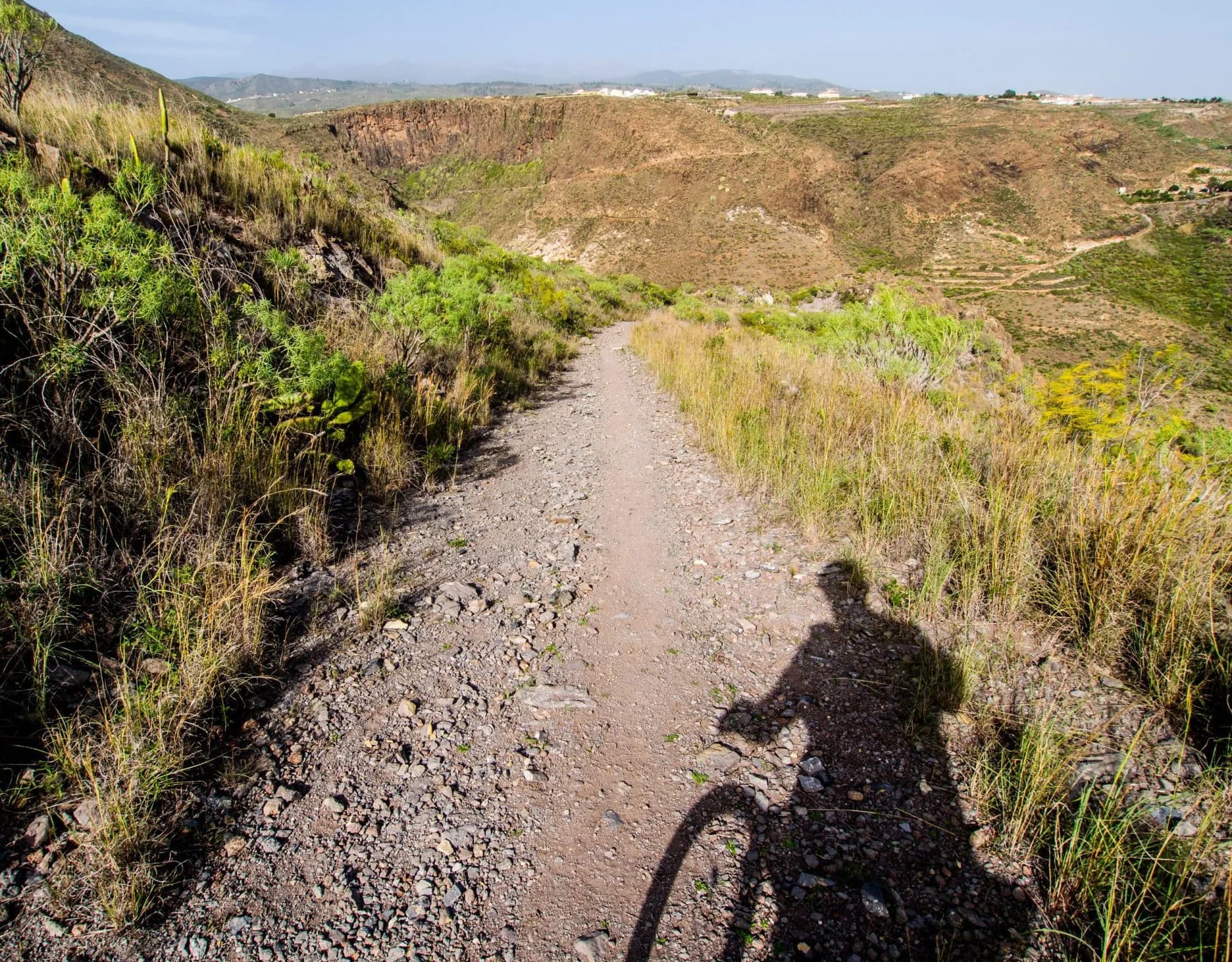 Cyclist's shadow on rocky dirt path through arid hills with distant houses