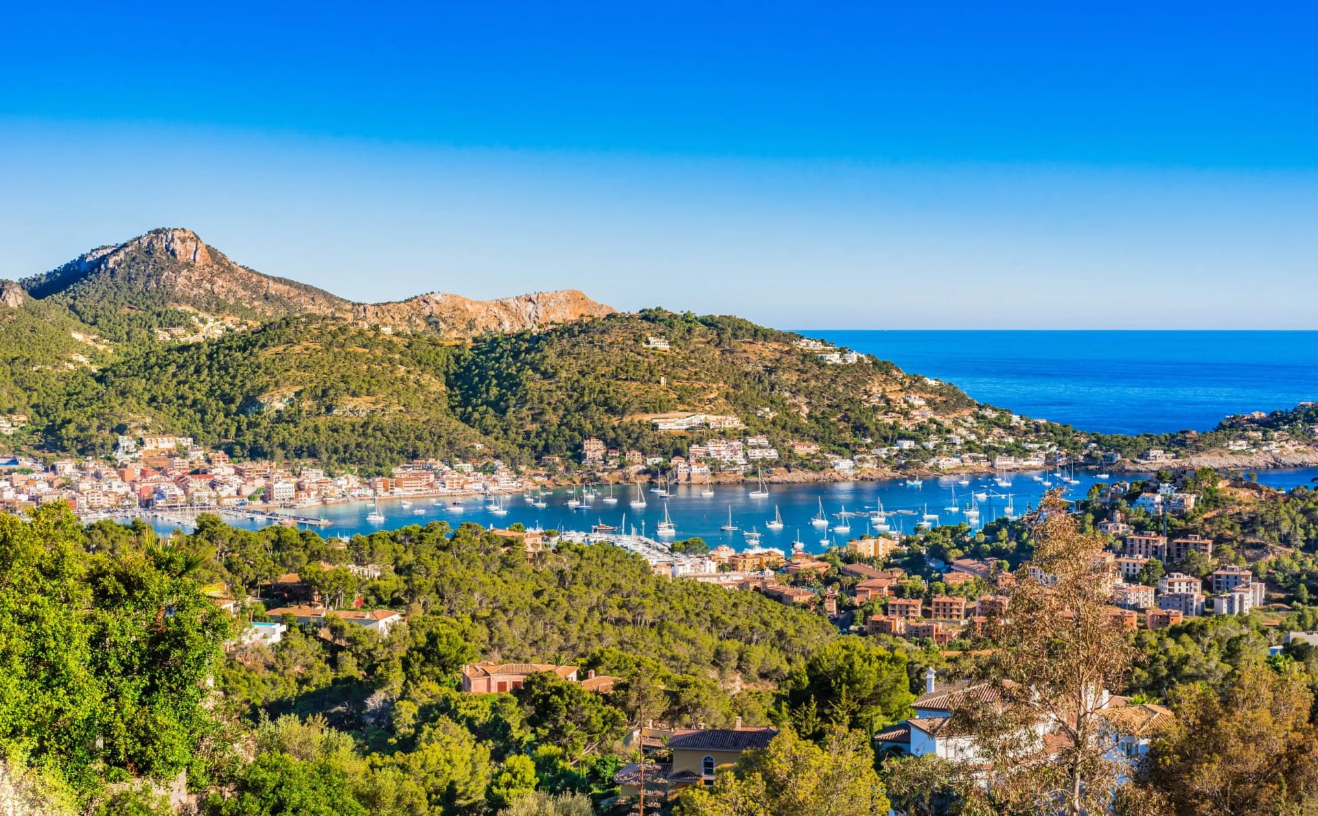 Coastal town nestled in green hills overlooking a bay filled with anchored sailboats in Andratx.