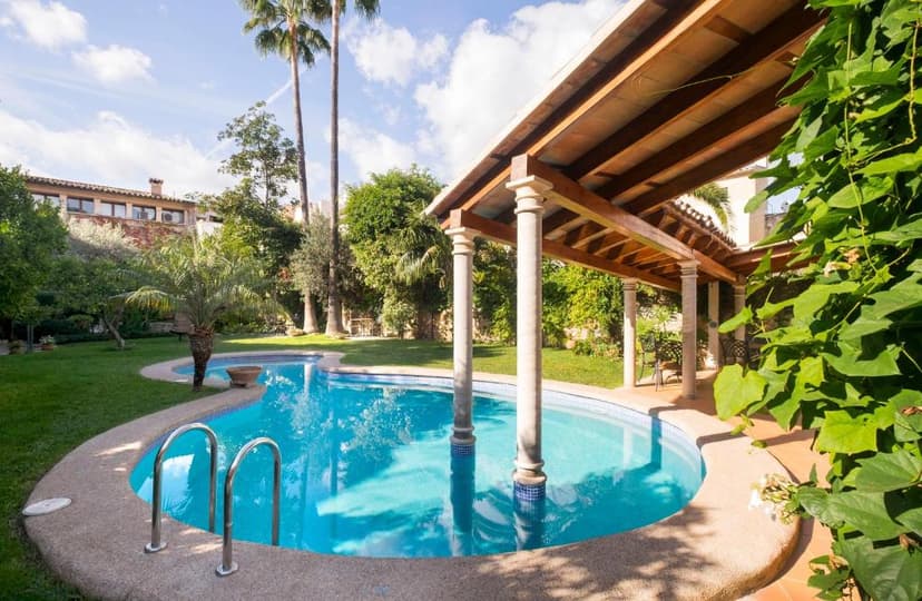 Swimming pool with curved edge, covered pergola, and lush green garden with palm trees.