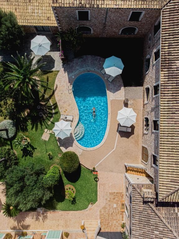 Aerial view of stone hotel courtyard with swimming pool, sun umbrellas, and lush green garden.