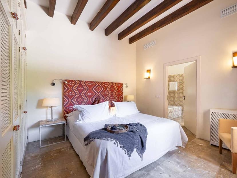 Bedroom with white bedding, patterned headboard, exposed wooden beams, and view into bathroom.