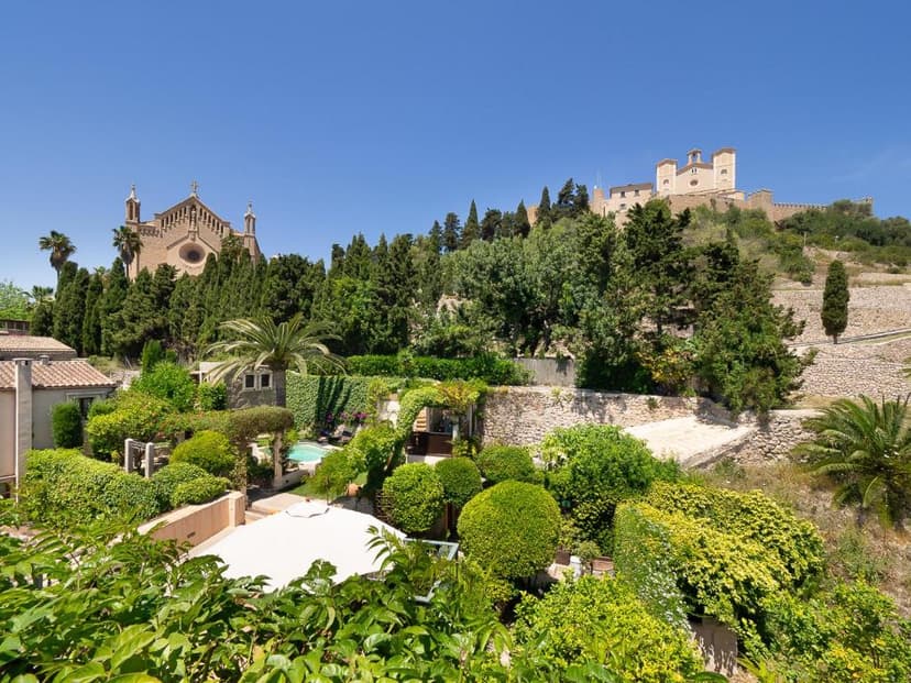 Lush gardens with pool overlook a church and castle on a sunny hillside, likely in Artà.
