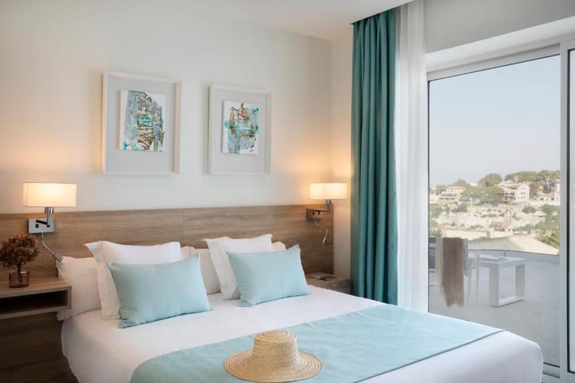 Bedroom with white and blue bedding, straw hat, and balcony view of coastal hillside buildings.