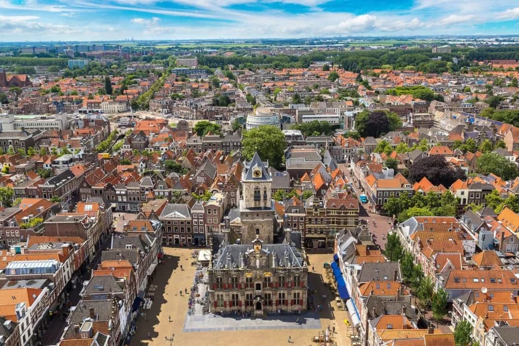 Aerial view of Delft city center with historic town hall and dense orange rooftops under blue sky.