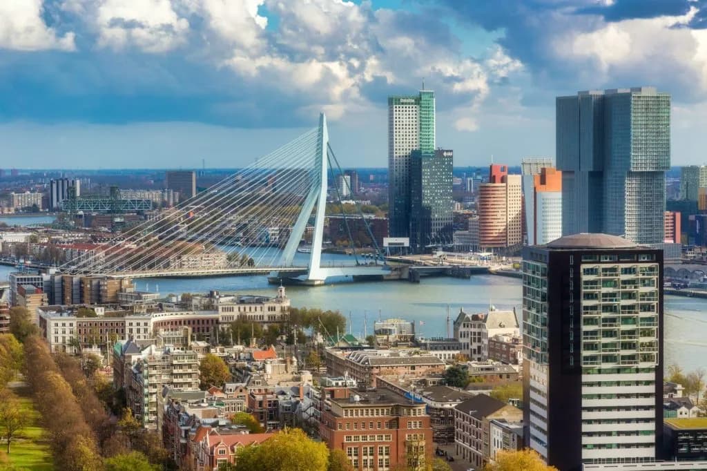 Rotterdam cityscape with Erasmus Bridge, modern skyscrapers, and dense buildings under a cloudy sky.
