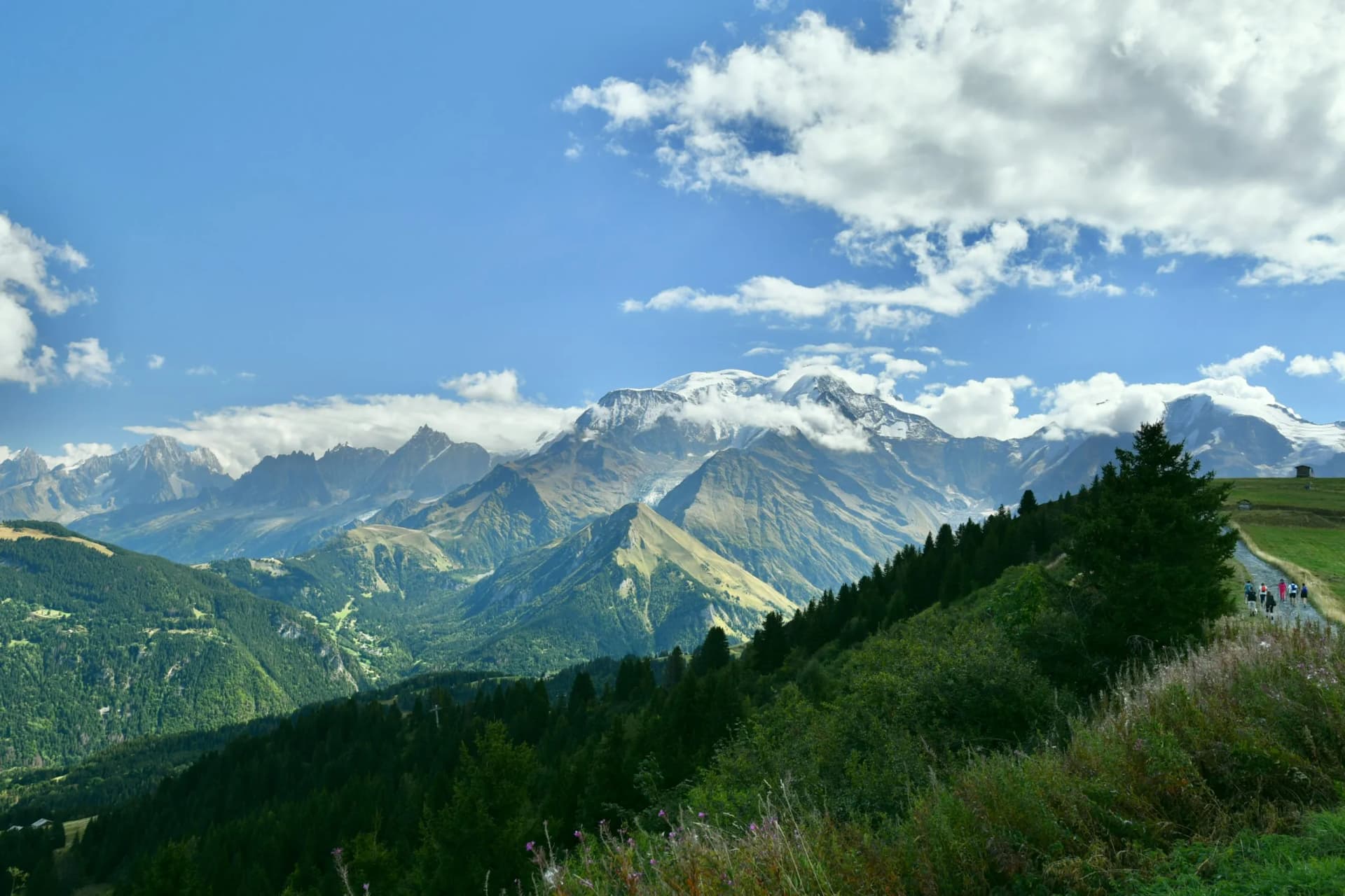 Hiking on trail toward Mont Blanc massif with snow-capped peaks under blue sky.