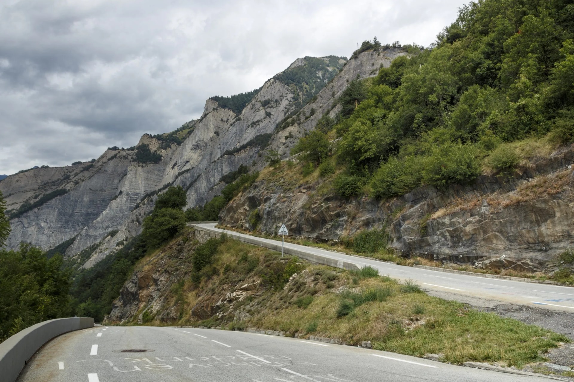 Winding mountain road ascent near steep, layered gray cliffs and green foliage under cloudy skies in La Grave, France.