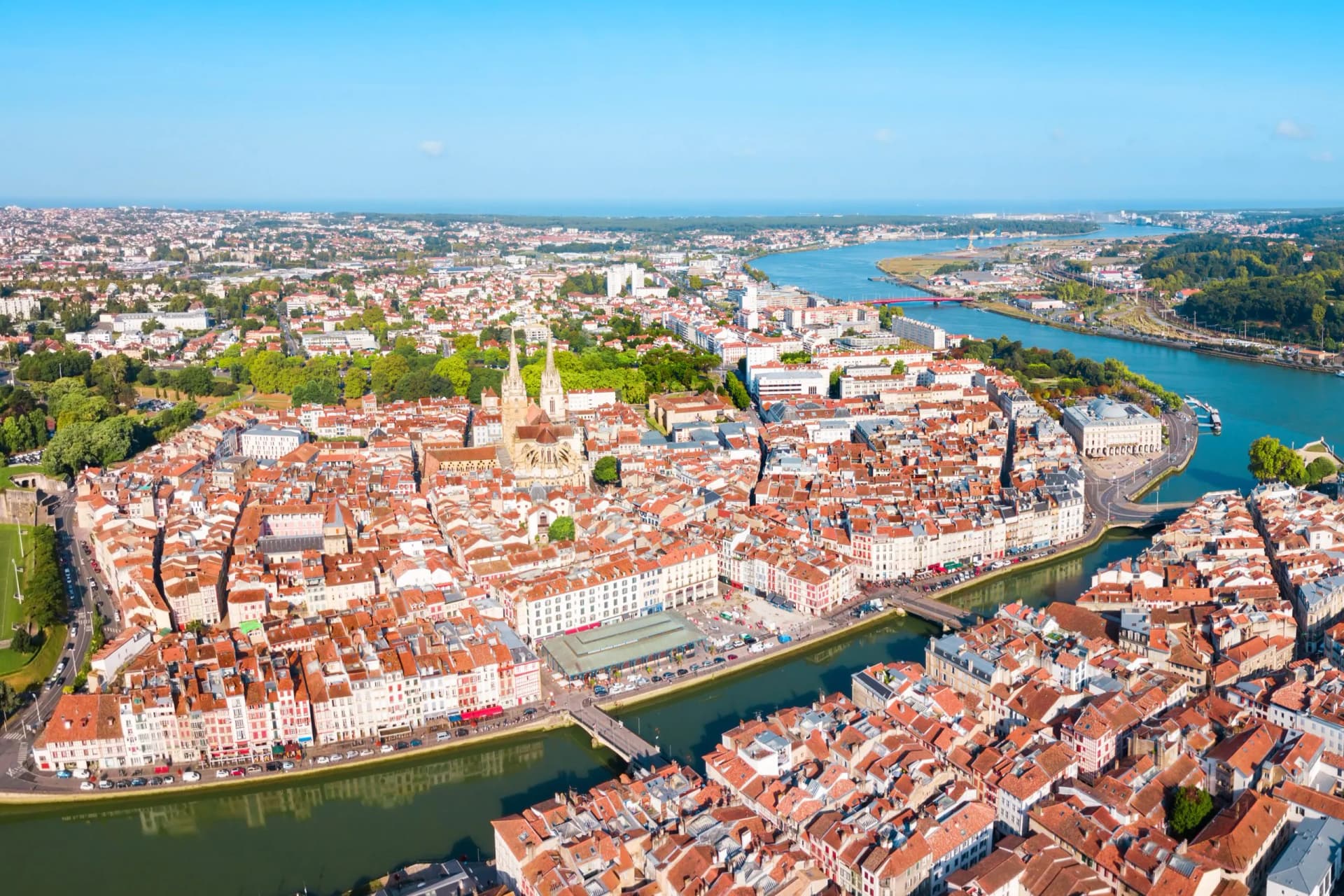 Aerial panoramic view of Bayonne city with terracotta roofs, a river, and the ocean in the distance.