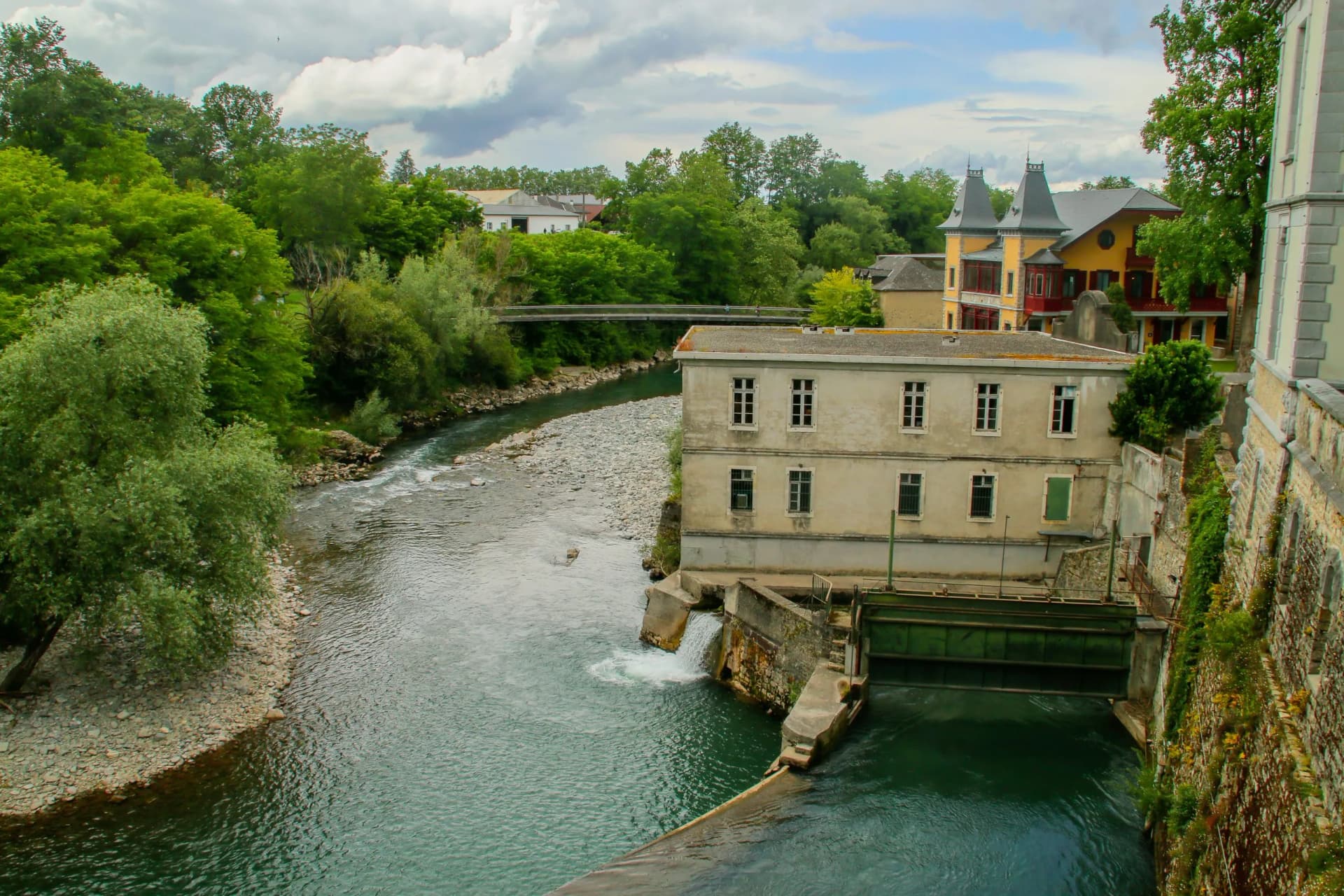 River flowing past stone buildings with a small dam and lush green trees under a cloudy sky.