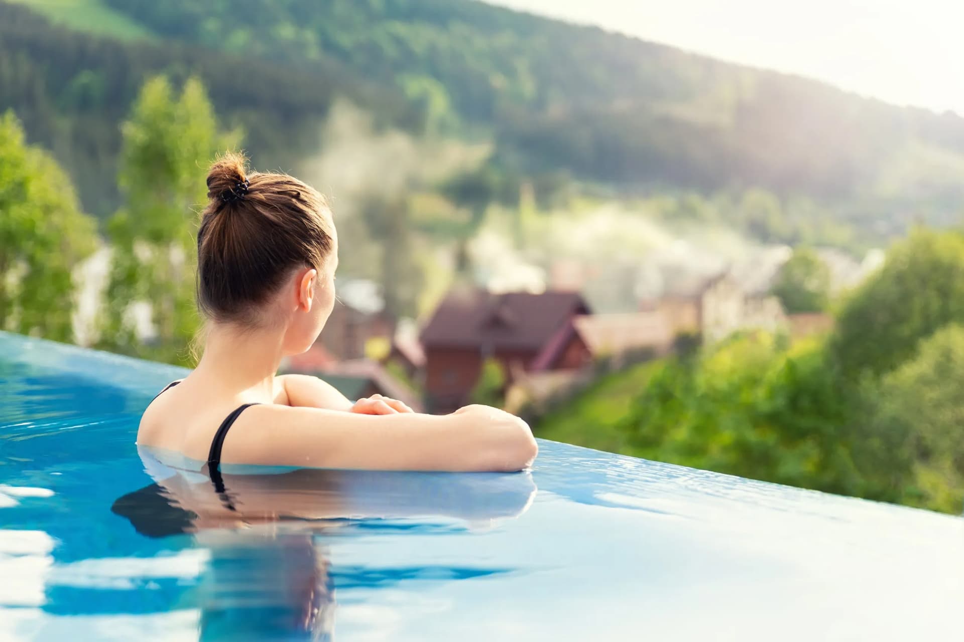 Woman relaxing in infinity pool overlooking foggy green mountains at sunset in alpine wellness resort.
