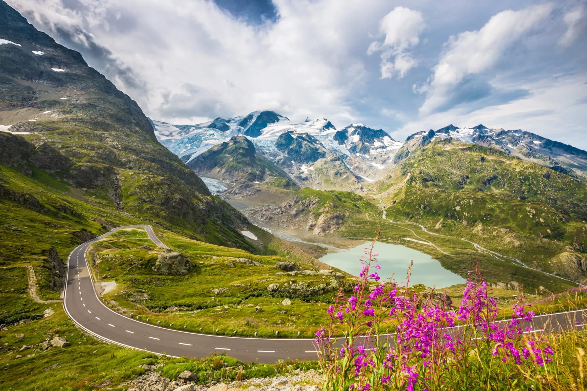 Winding mountain pass road in the Alps with glacier peaks, lake, and purple wildflowers in summer.