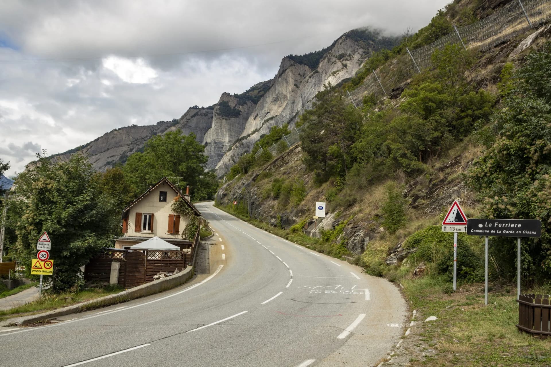 Winding mountain road ascent near La Ferriere, France, with steep cliffs and small house.