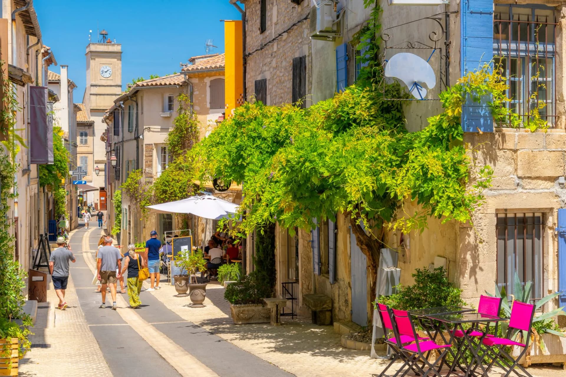 Pedestrians walk up a sunny street toward a clock tower in Saint-Remy-de-Provence.
