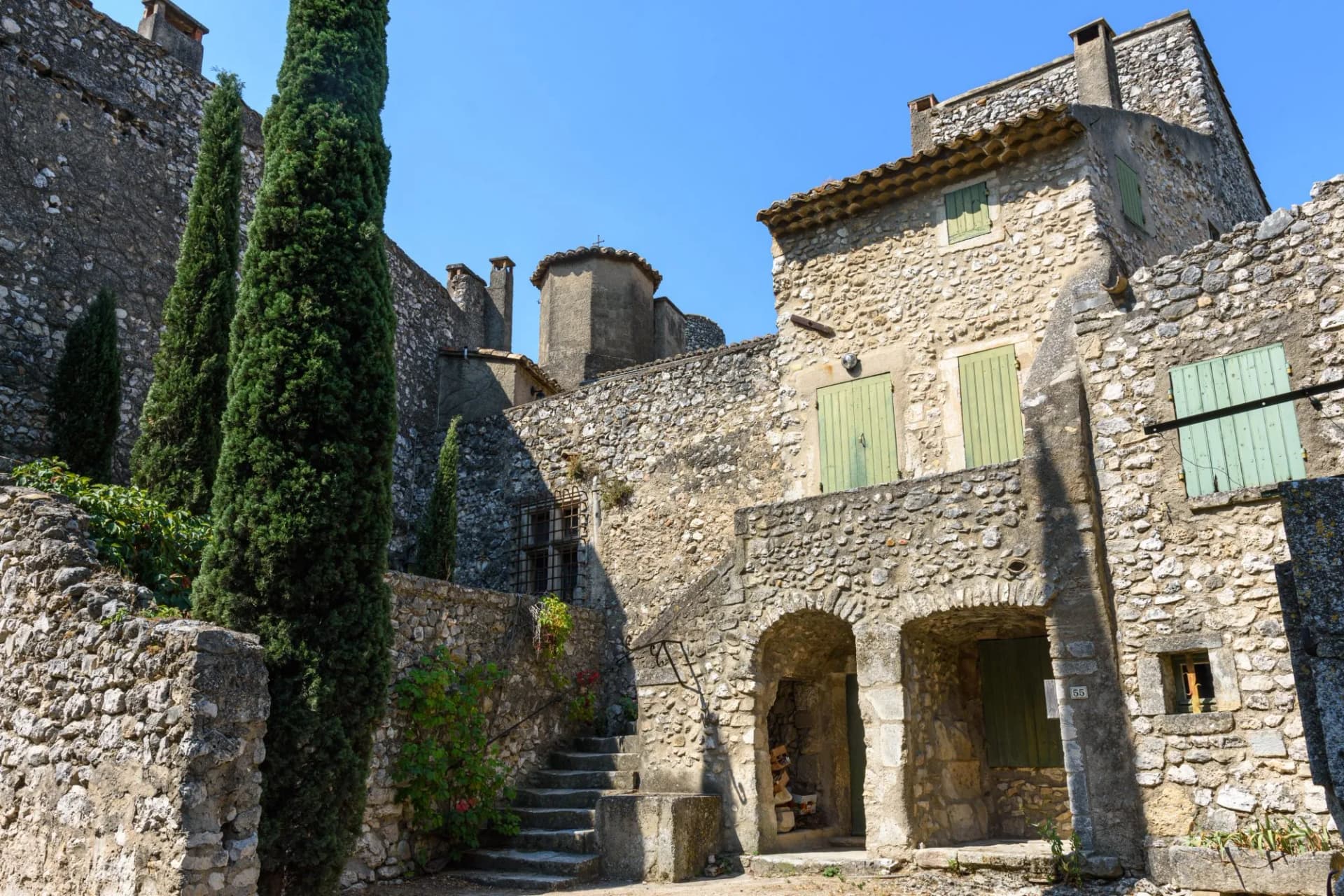 Stone buildings with green shutters and tall cypress trees in Eygalières under a clear blue sky.