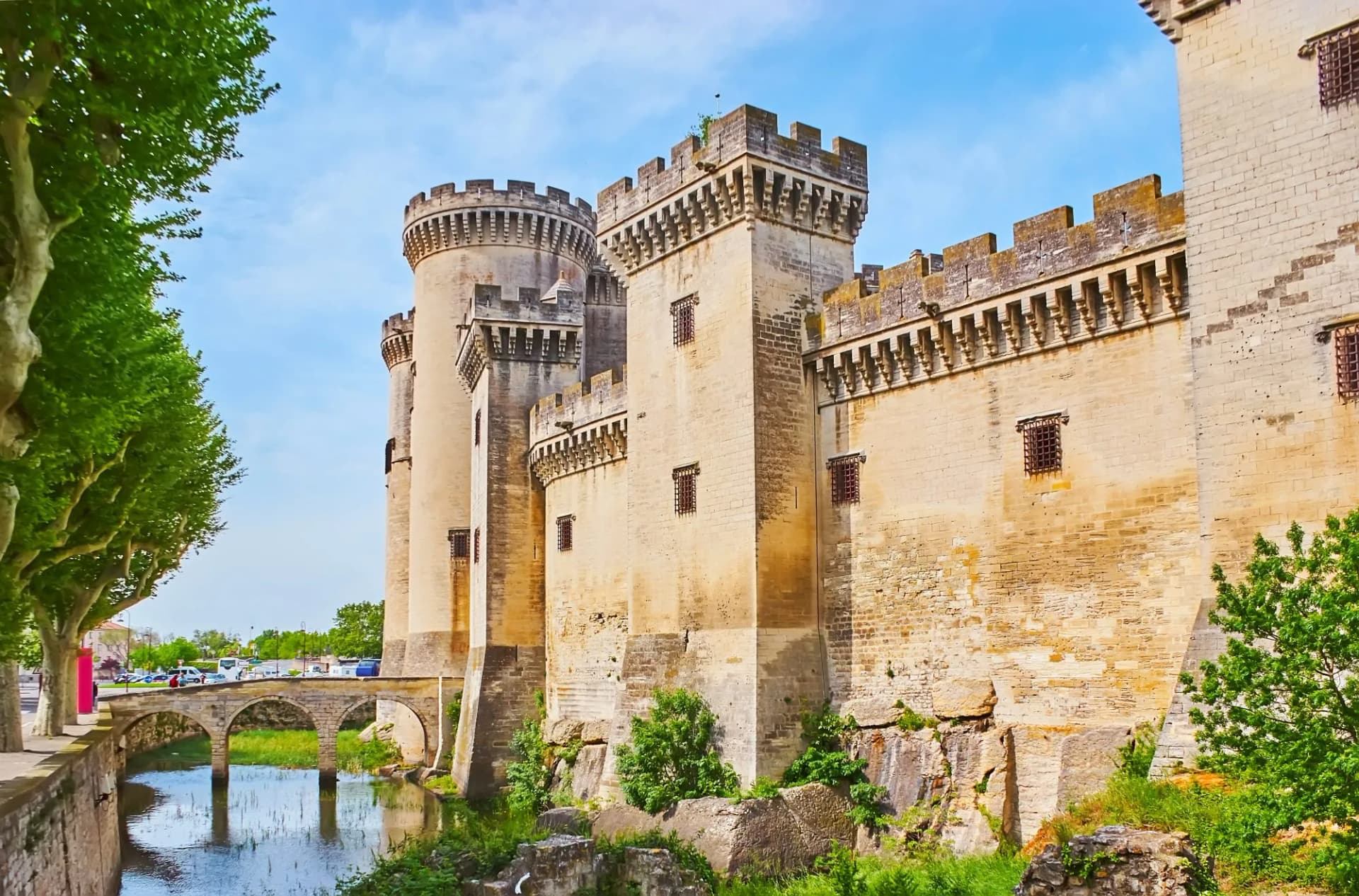 Tarascon Castle stone towers next to a moat and arched bridge under a blue sky.