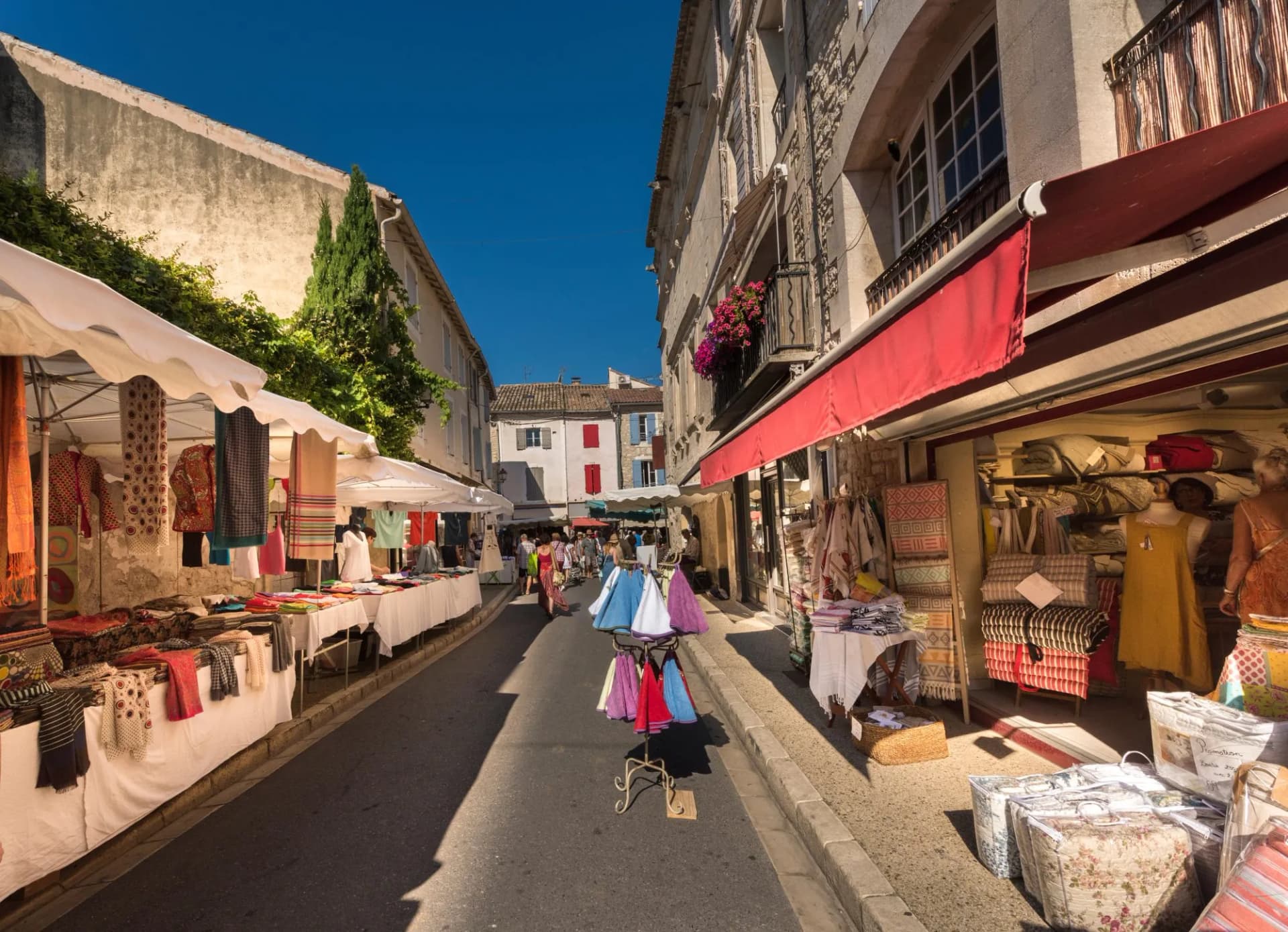 Market stalls with textiles line a narrow street in Saint-Remy-de-Provence under a bright blue sky.