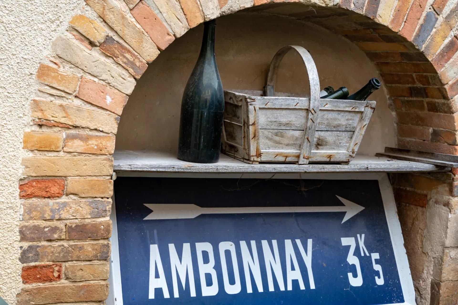 Ambonnay direction sign under a brick arch with a wine bottle and wooden basket.