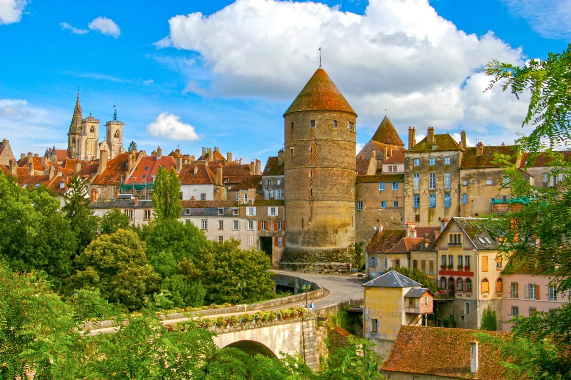 Historic town of Beaune featuring a large round tower, stone bridge, and church spires under a blue sky.