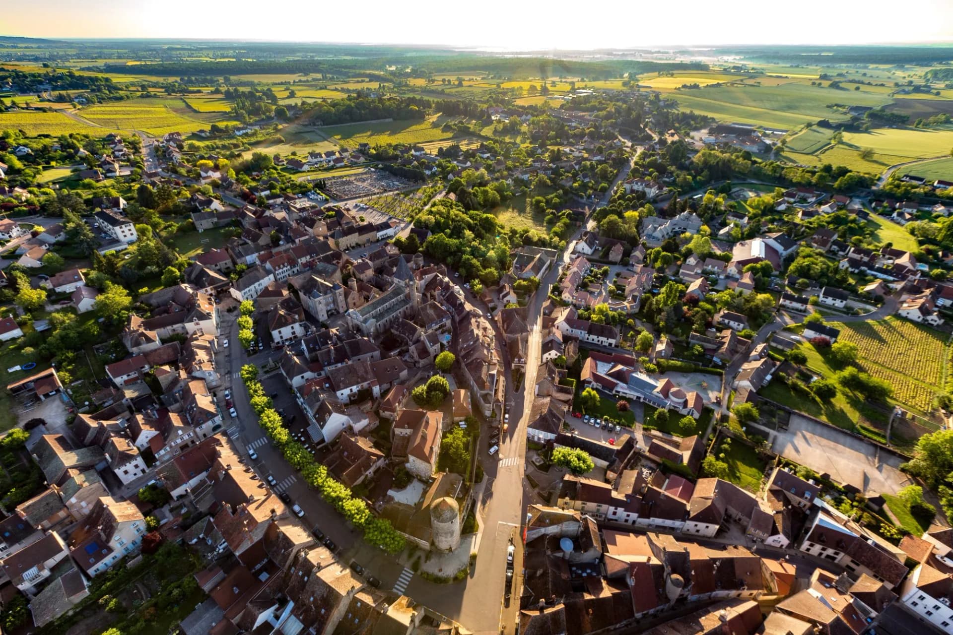 Aerial view of historic European village with terracotta roofs surrounded by green fields and vineyards at sunrise.