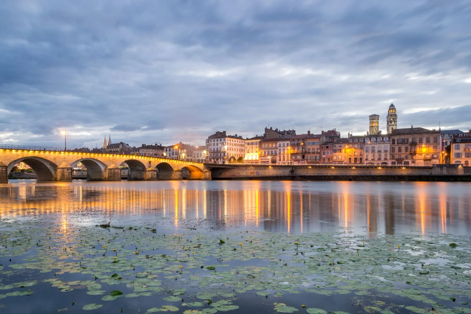 Stone arch bridge over river with illuminated city buildings and lily pads in Macon.