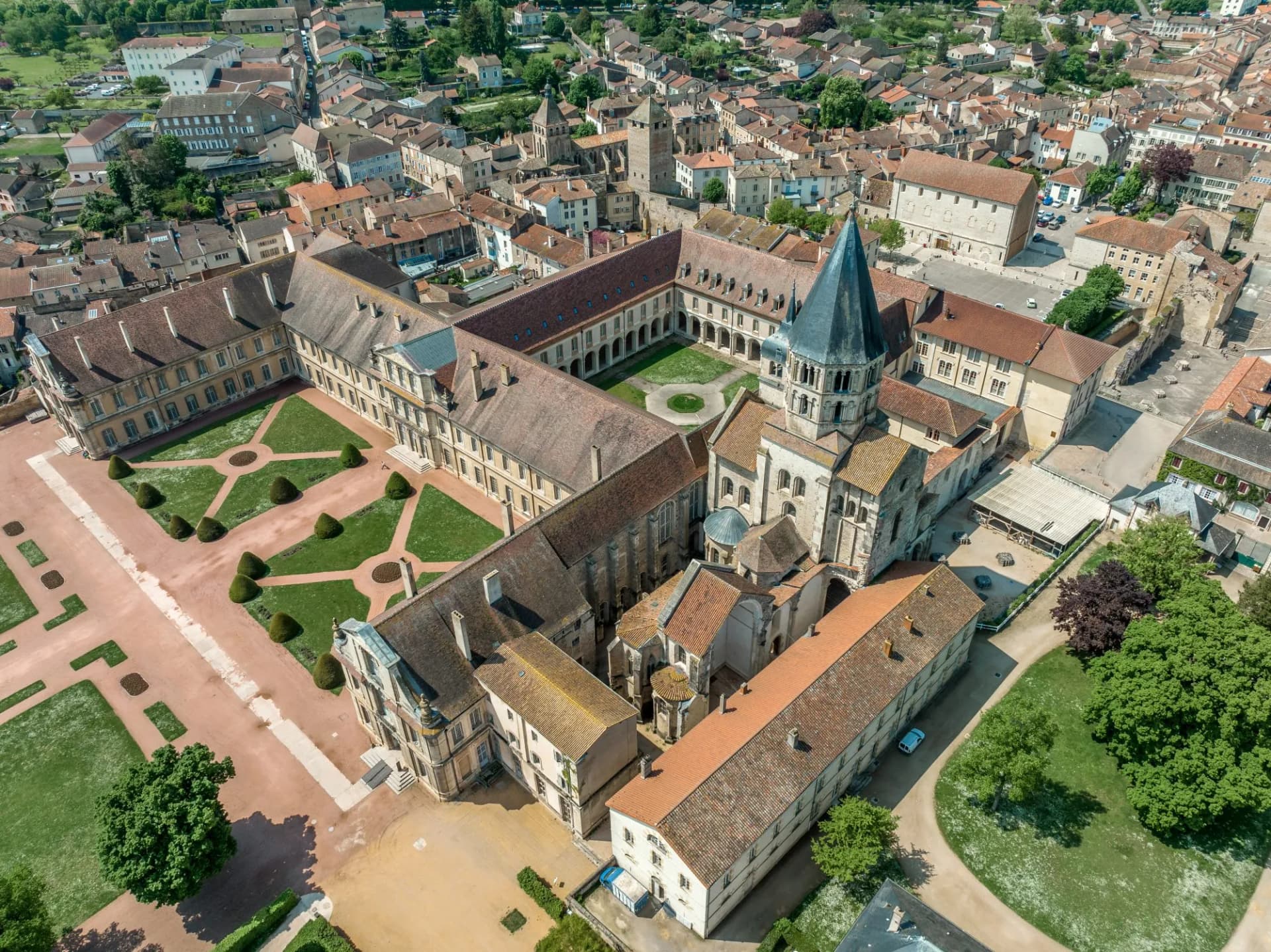 Aerial view of large historic abbey complex with conical spire and formal gardens in a European town.