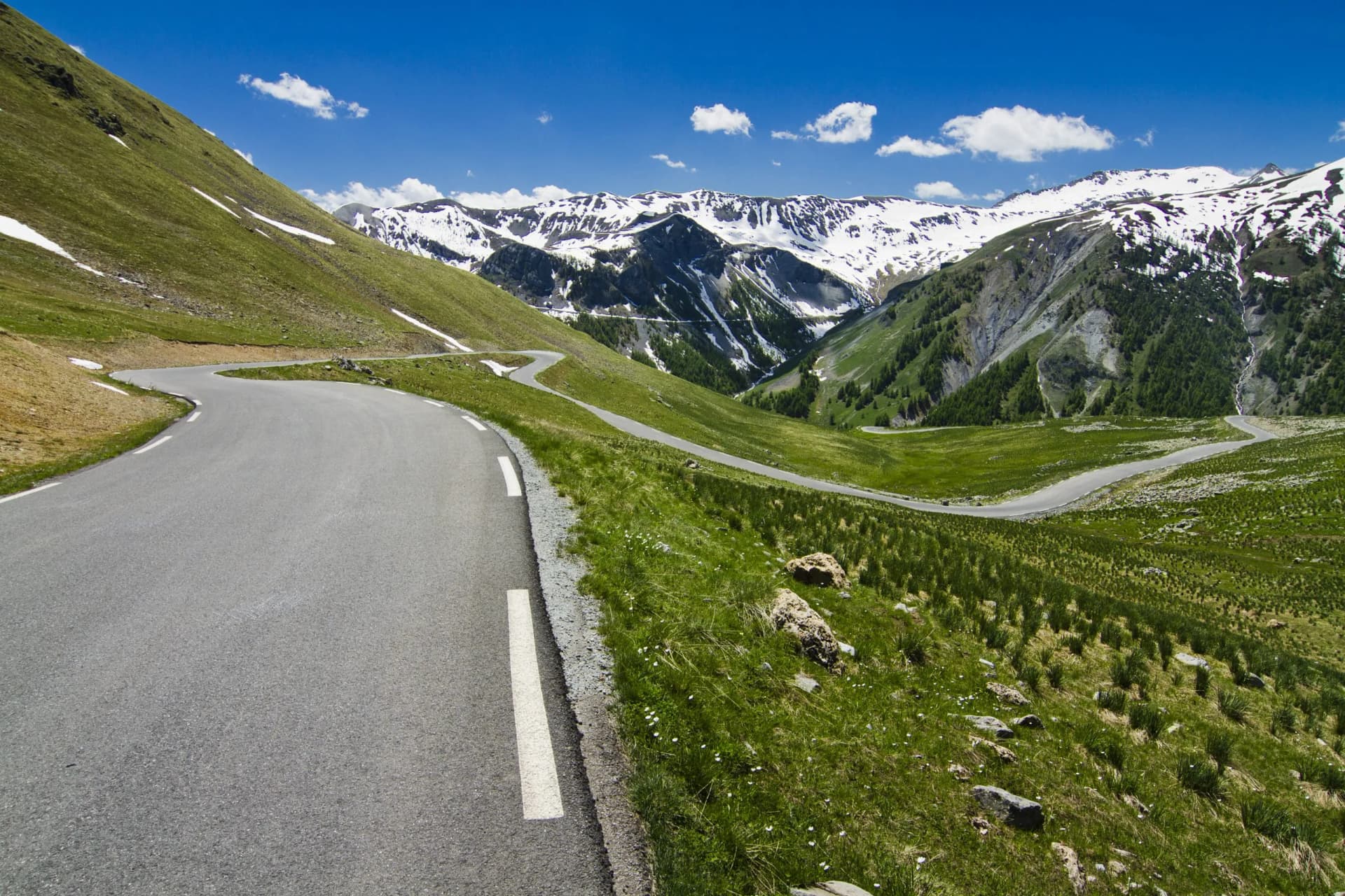 Winding mountain road ascending to Col de la Bonette with snow-capped peaks under a blue sky.