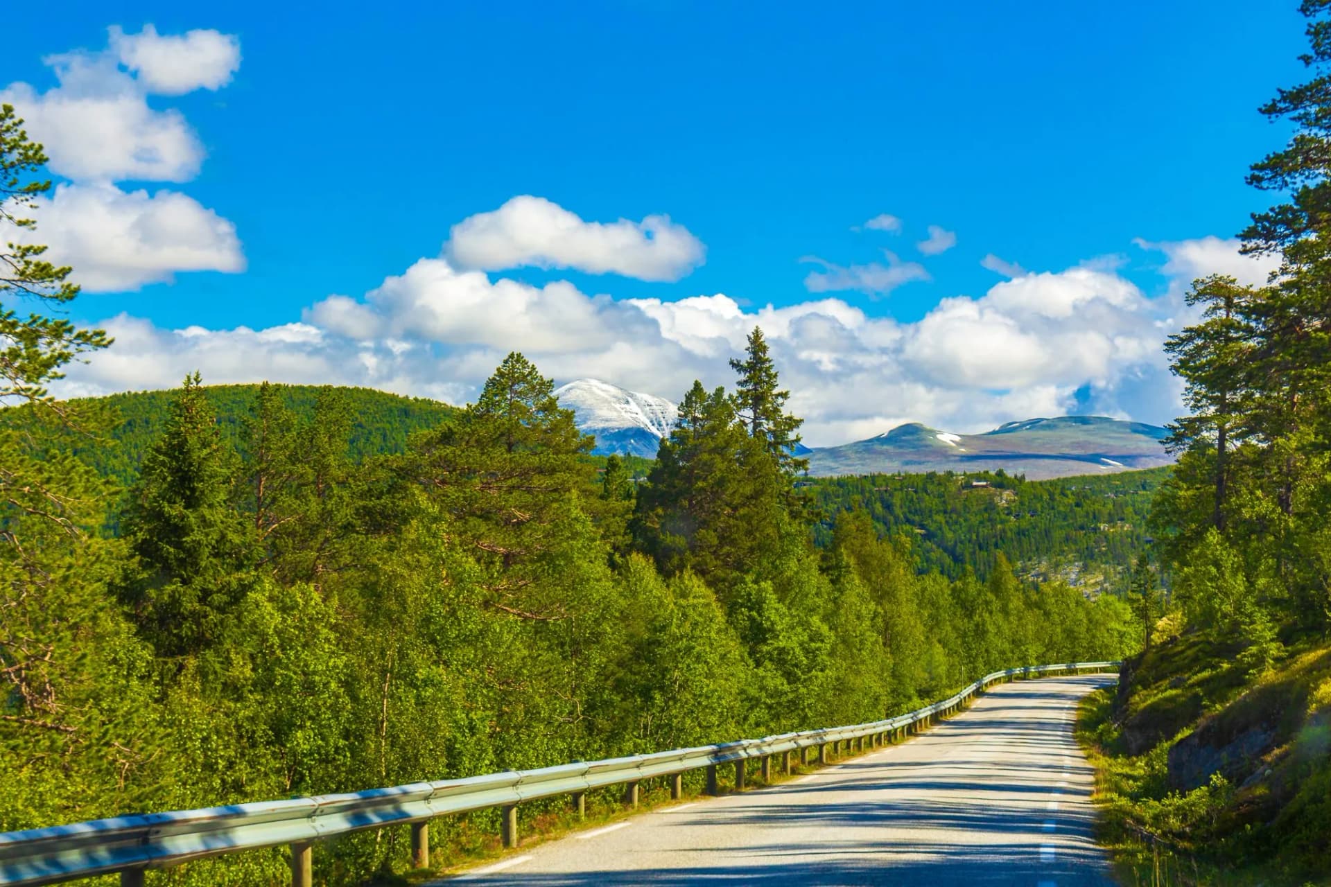 Mountain road curving through green forest with snow-capped peaks under blue sky in Kvitfjell.