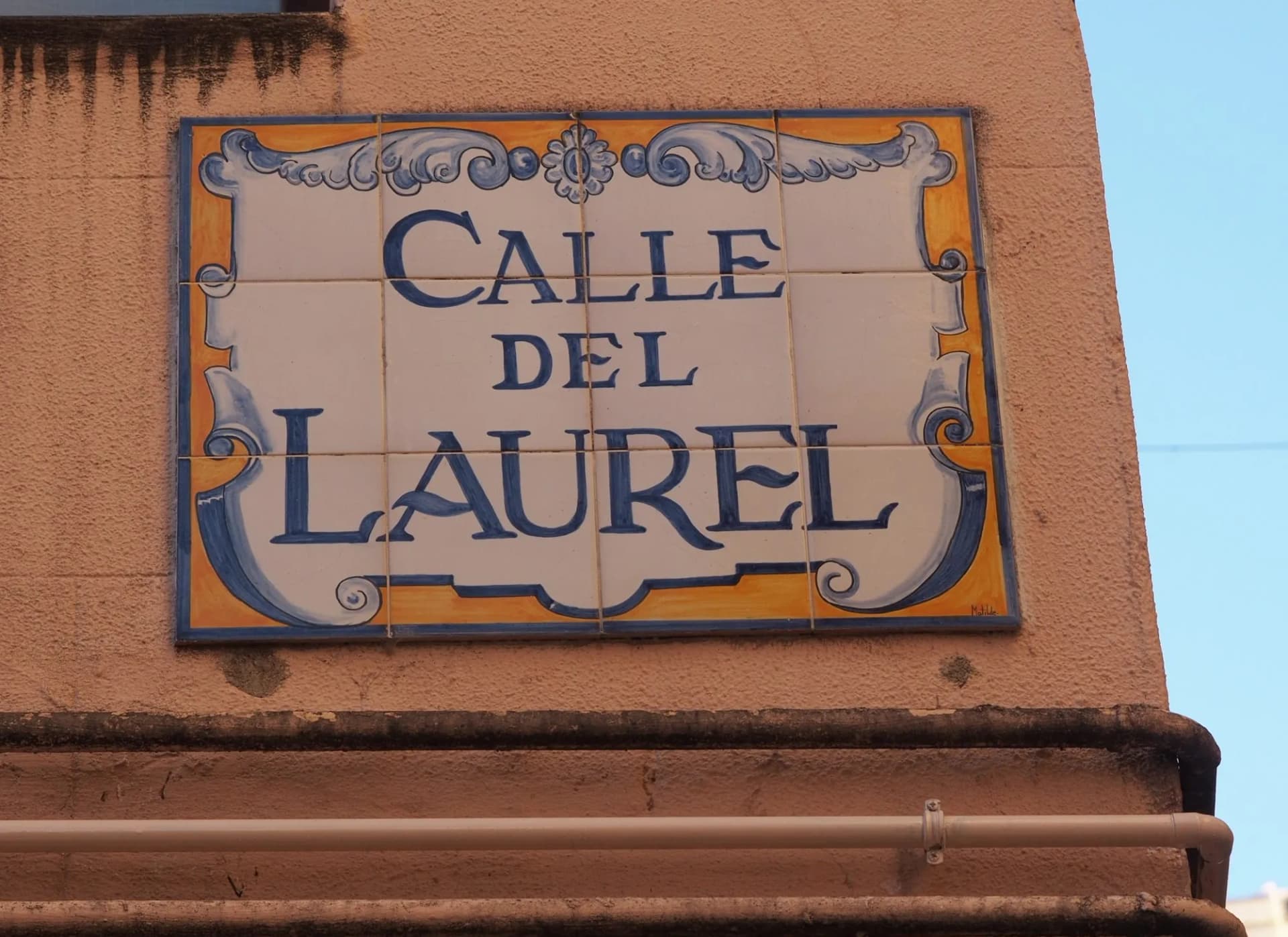 Ceramic street sign reading "CALLE DEL LAUREL" on textured peach wall under blue sky.