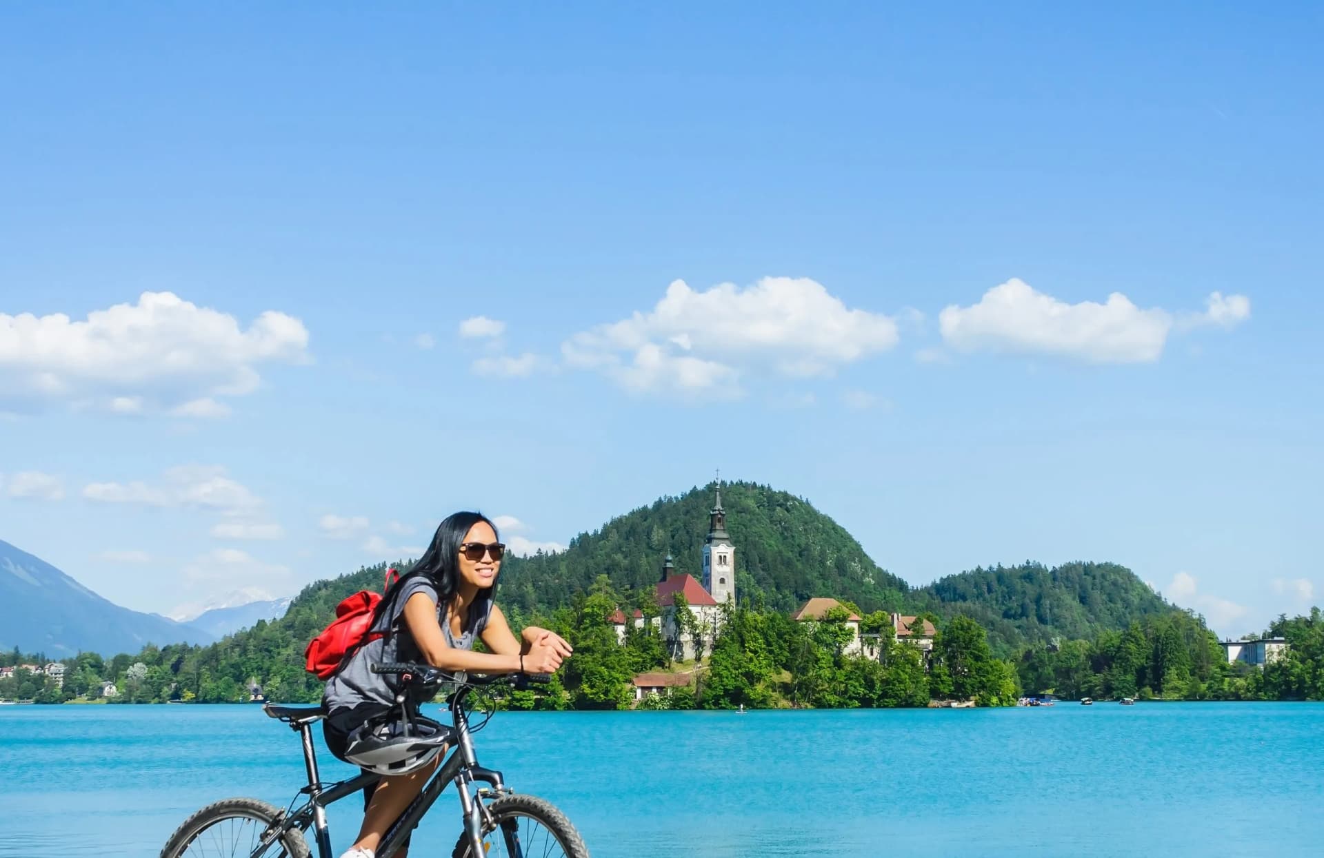 Cyclist with backpack resting by turquoise Lake Bled with island church and mountains.