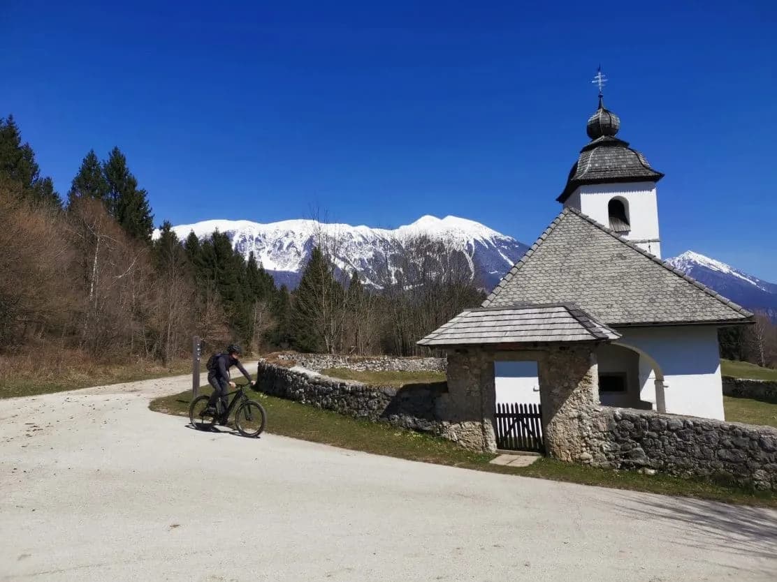 Mountain biker rides past St. Katarina Church above Bled with snow-capped mountains.