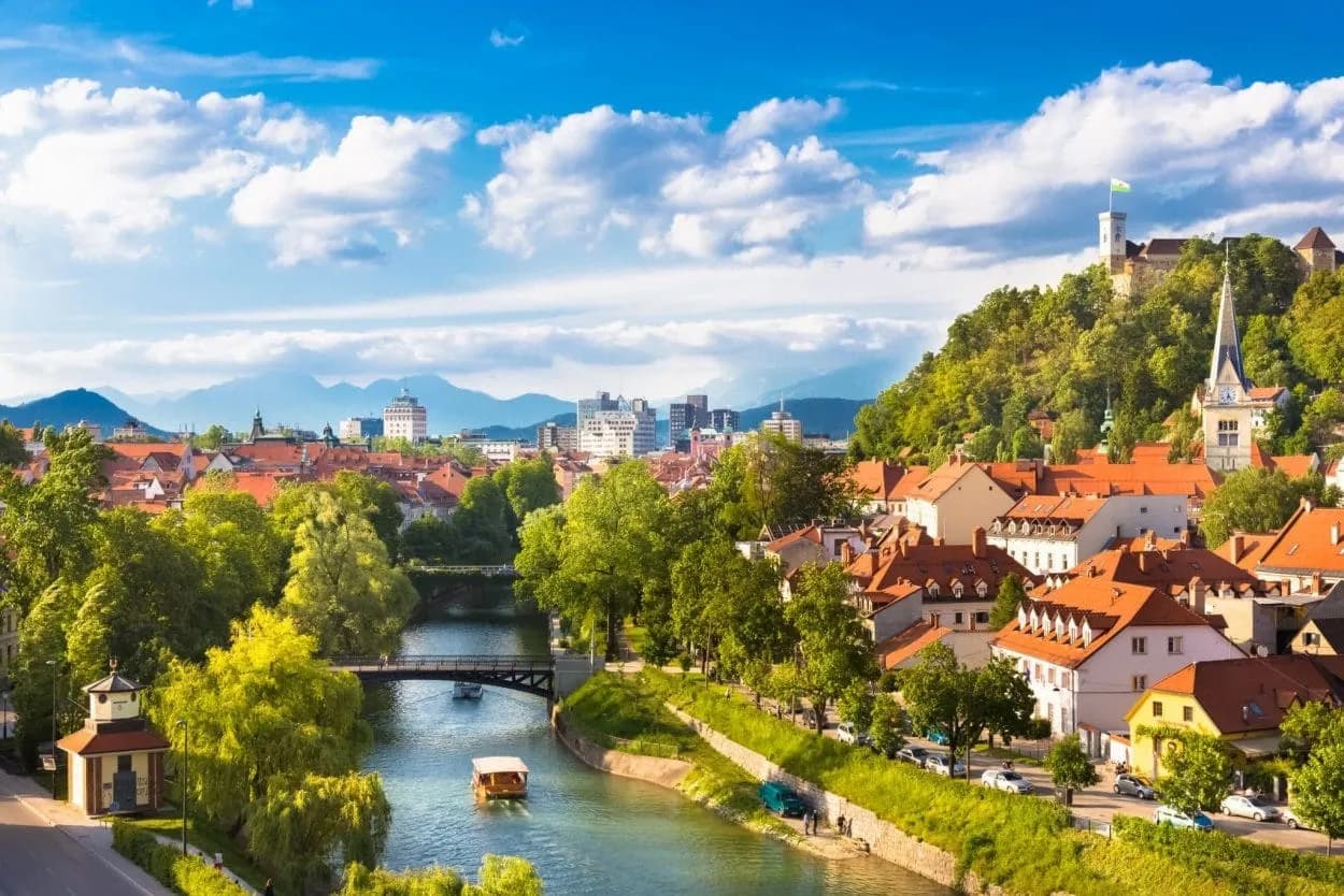 Ljubljana city view with river boats, Ljubljana Castle on a green hill, and mountains in the distance.