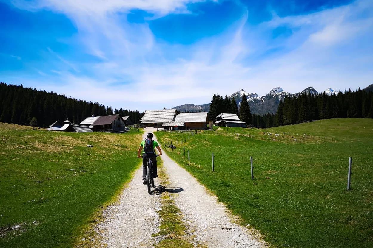 Cyclist on gravel path toward wooden huts with snowy mountains in Pokljuka, Zajavornik.
