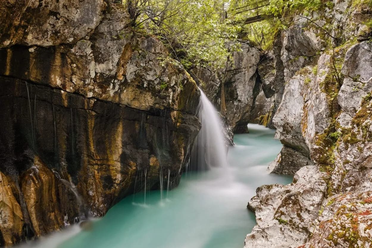 Waterfall cascading into turquoise river flowing through steep, mossy rock gorge with lush greenery.