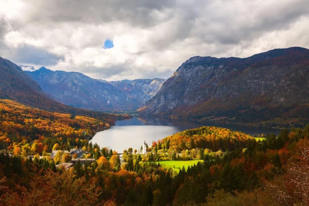 Lake Bohinj in Triglav National Park with autumn foliage and dramatic mountains under cloudy sky.