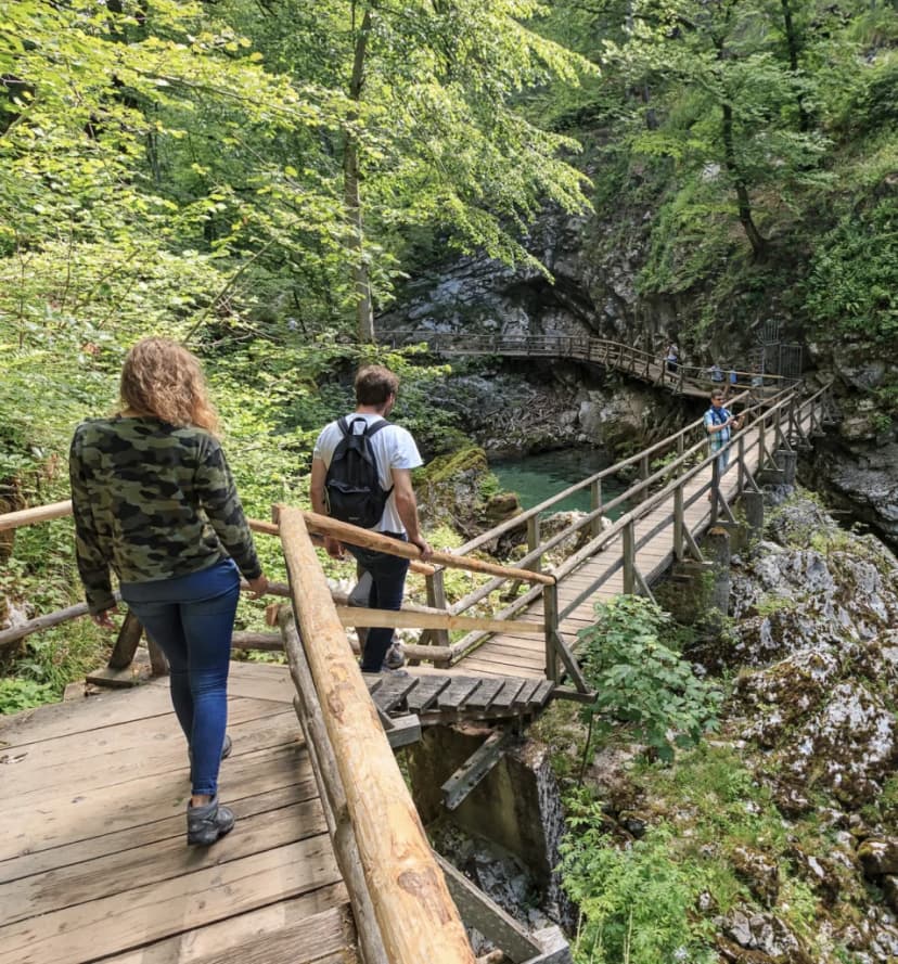 Hikers walking on wooden boardwalks over a turquoise river gorge in a lush forest.