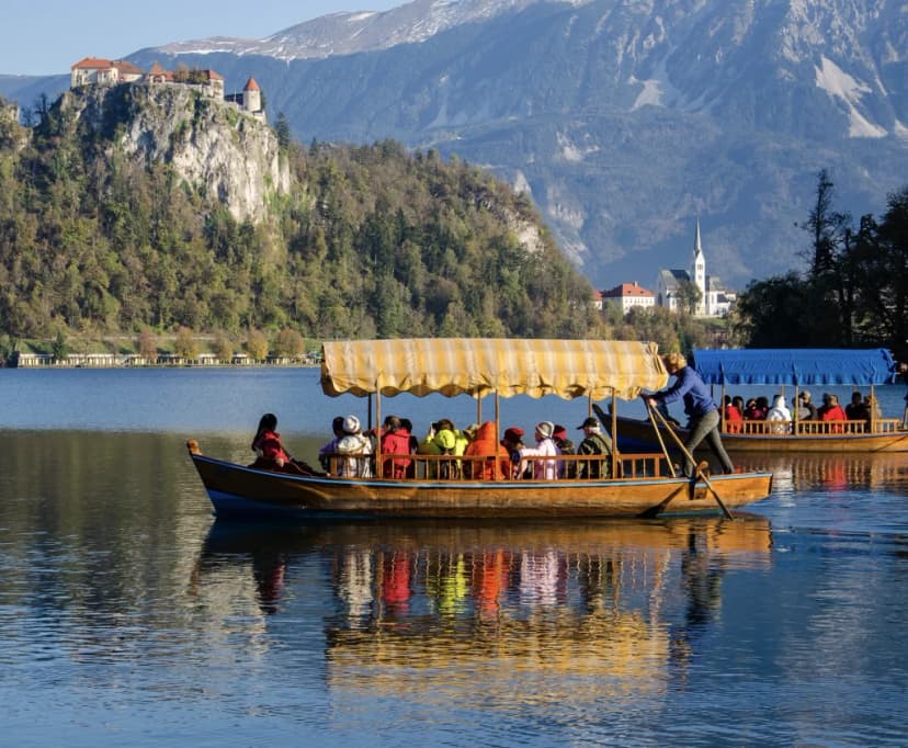 Pletna boats carrying tourists on Lake Bled with Bled Castle on cliff and mountains.