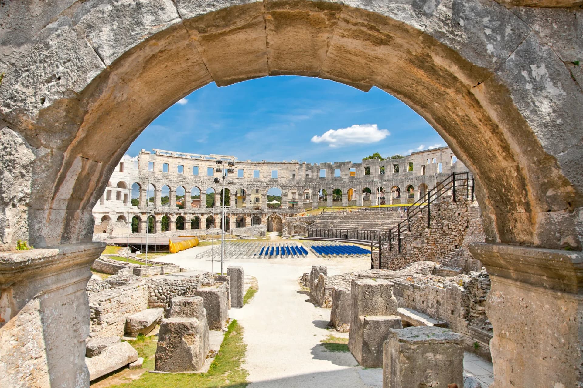 Roman Amphitheatre Arena in Pula viewed through a large stone archway on a sunny day.