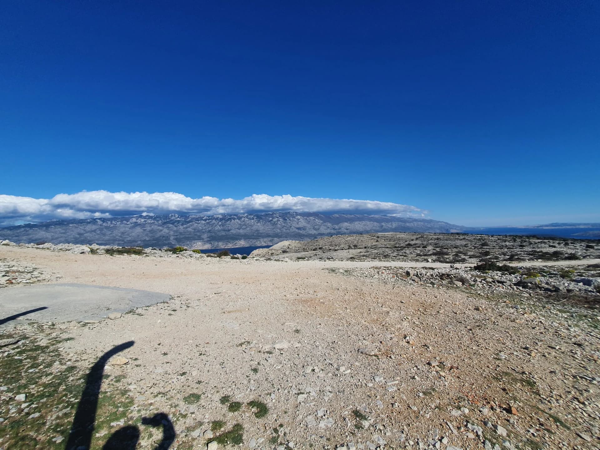 Rocky, arid landscape overlooking the sea with distant snow-capped mountains under blue sky.