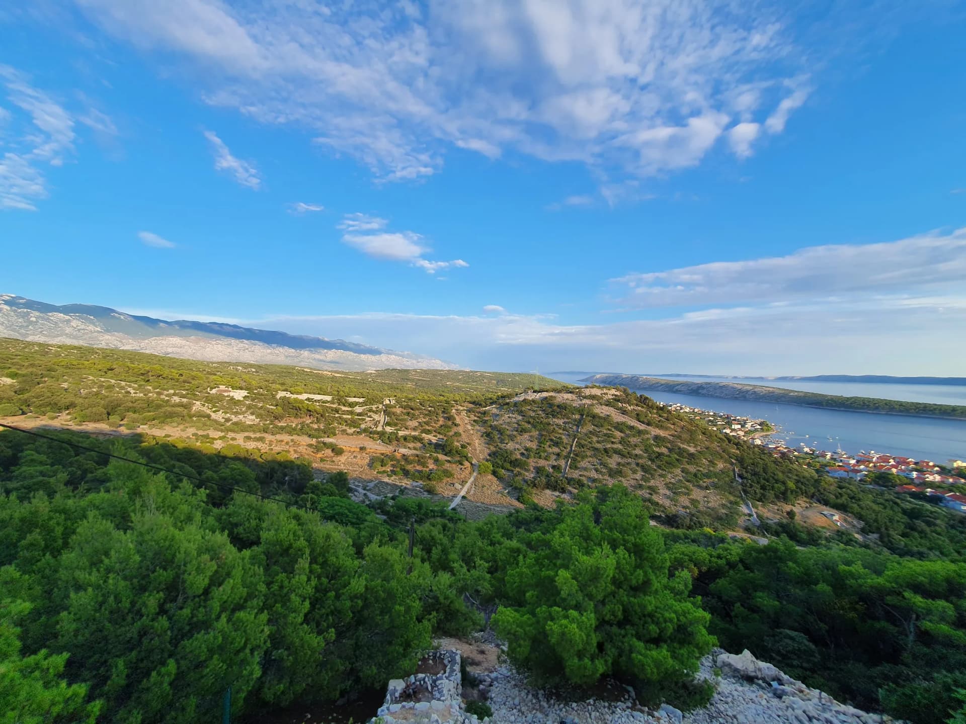 Hilly coastal landscape with green trees, town, and mountains under a blue sky.